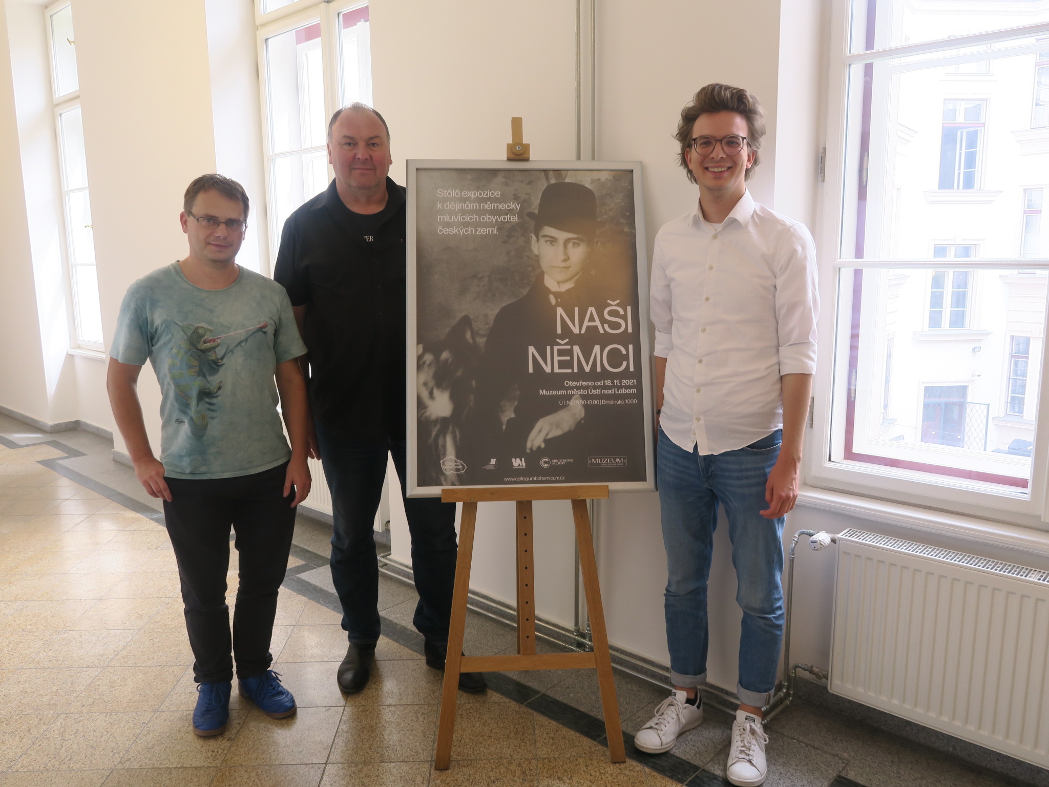 In der Mitte steht ein Plakat der Ausstellung in einem Gang. Rechts davon steht Leon Eckert. Links steht Erhard Grundl. Daneben steht die Person, die die Museumsführung gemacht hat.